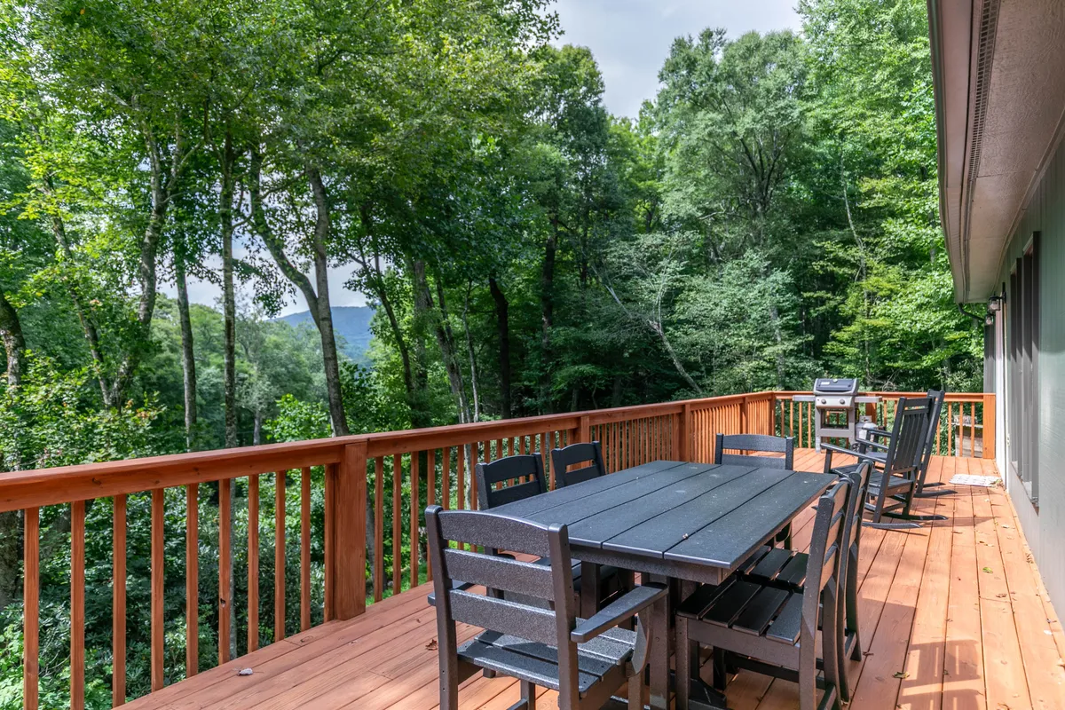 A tranquil wooden deck furnished with a dining table and grill sits amidst leafy trees, inviting outdoor dining, barbecues, and peaceful moments in nature. The setup is open, accessible, and designed for communal enjoyment.