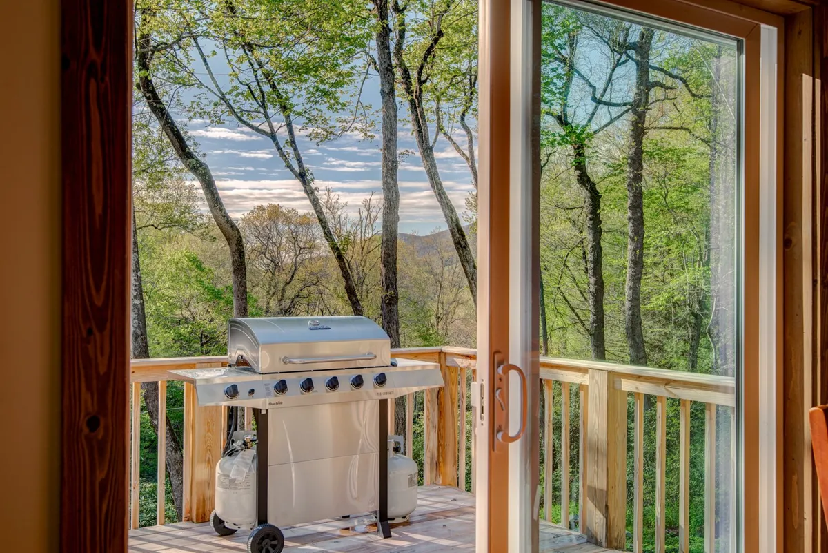 A spacious wooden deck framed by lush green trees and an open sky, featuring a grill and comfortable seating. The deck overlooks the natural landscape, inviting guests to relax, dine, or entertain while fully immersed in the peaceful outdoors.