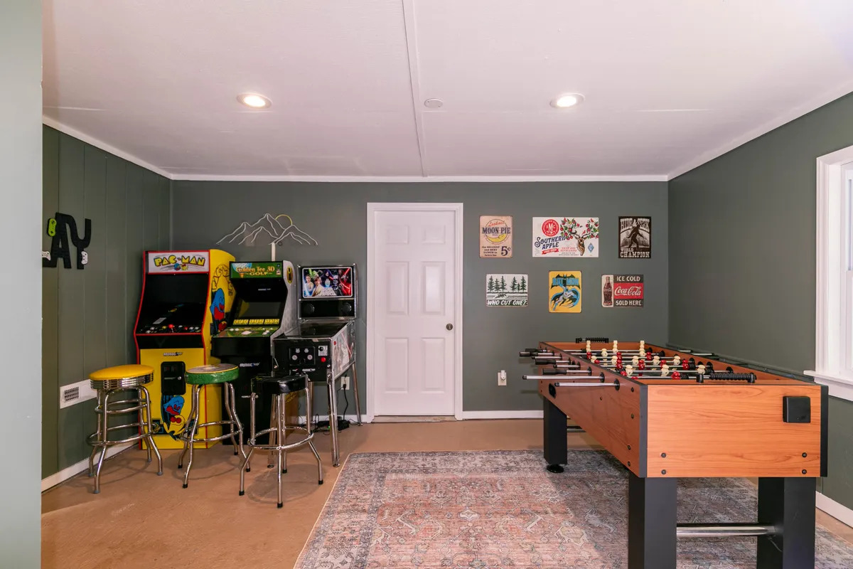 A vibrant game room featuring a foosball table in the foreground, classic arcade consoles including Pac-Man and Star Wars pinball, colorful vintage posters on the walls, and bright stools inviting groups to gather and play.