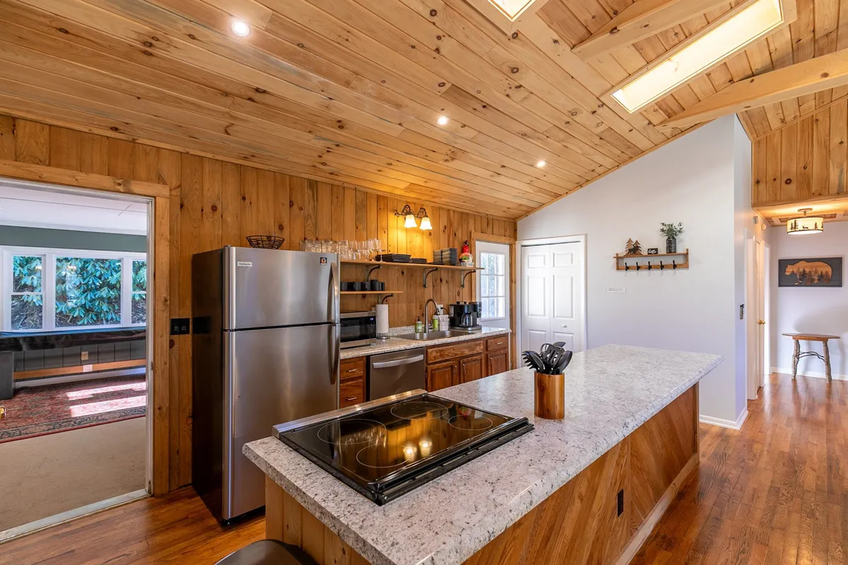 A close-up of the modern kitchen featuring a large kitchen island, stainless steel refrigerator, sleek granite countertops, and warm wooden cabinetry. The space looks newly updated and designed for both cooking and gathering, with a welcoming, accessible layout.