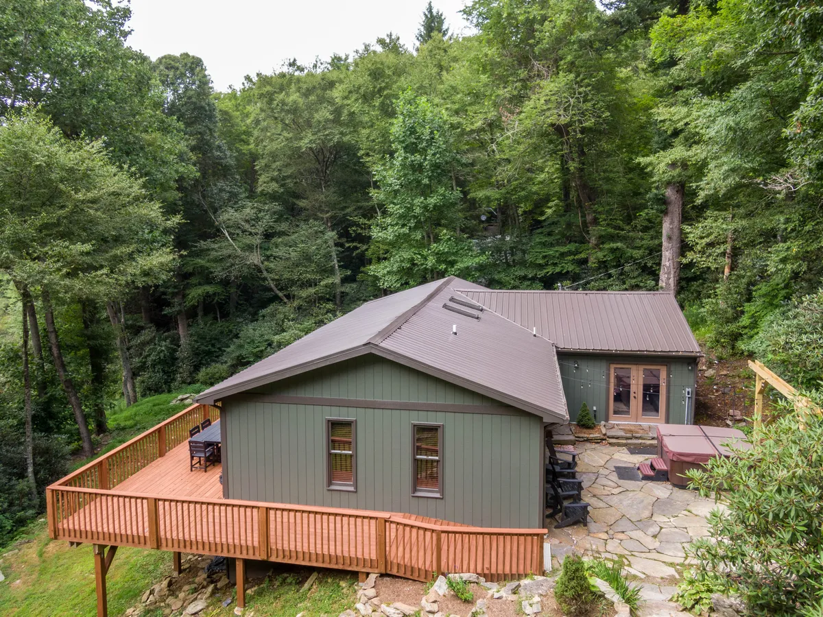 A broad view of the home's wooden deck overlooking a beautifully landscaped yard framed by lush green trees and a winding stone pathway. The deck features areas ideal for outdoor dining, relaxing in the bubbling hot tub, and gathering around a cozy fire pit. The tranquil setting is perfect for family conversations and unwinding after days of adventure.