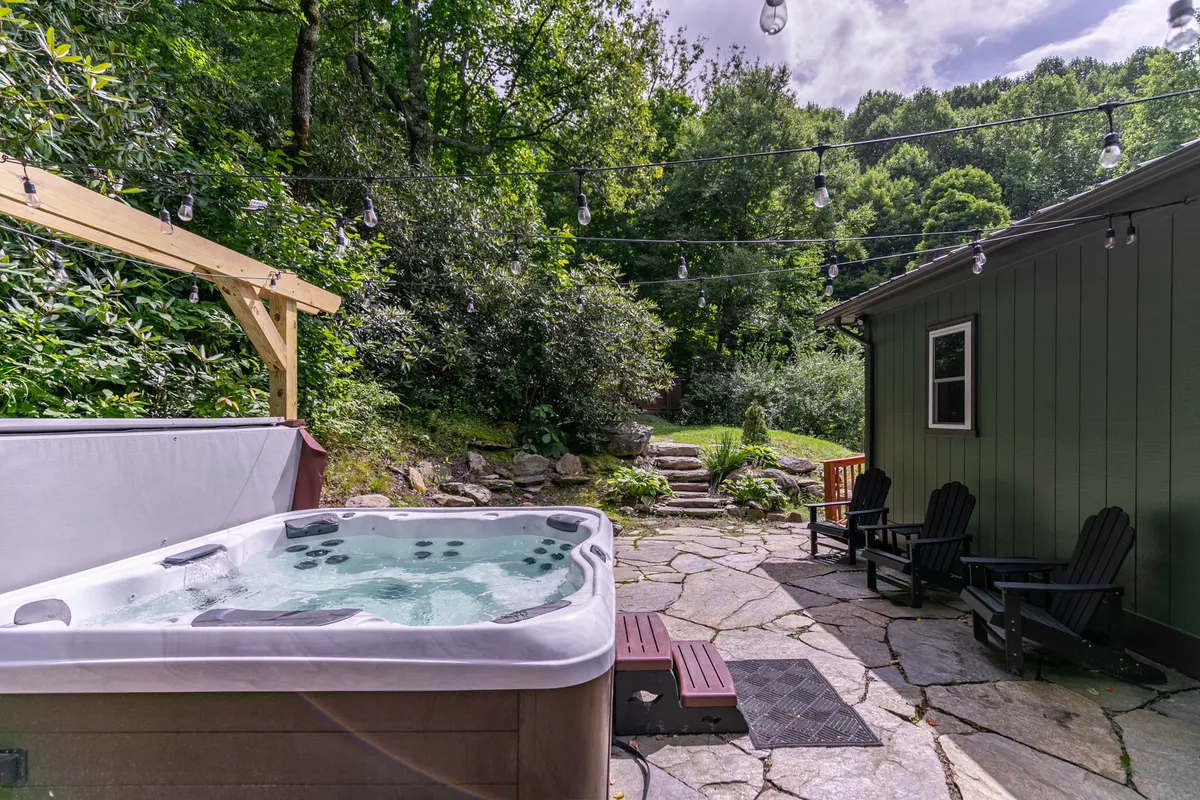 The scene presents a beautifully landscaped garden with a hot tub, string lights overhead, inviting outdoor seating, and a cozy fire pit area. The ambiance is warm and welcoming, perfect for evening relaxation or social gatherings under the stars.