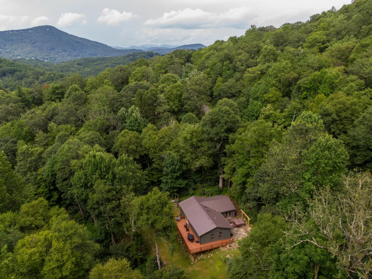 The image features the inviting exterior of a charming single-family house nestled among lush trees, with breathtaking views of the distant Blue Ridge Mountains. The setting captures the soft glow of sunset over the landscape, evoking a sense of peace and seclusion while still feeling accessible and welcoming. The expansive deck is visible, hinting at mornings spent with coffee and evenings under the stars.